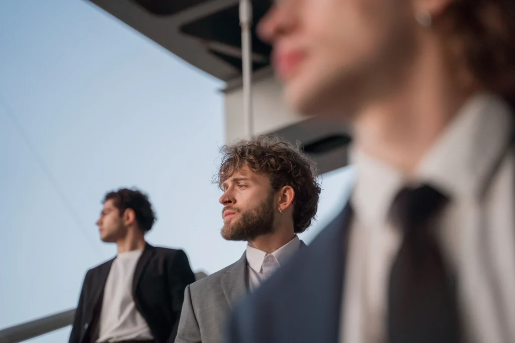 Non ci sono più i maschi di una volta. Per fortuna? Tre giovani uomini in giacca guardano in direzioni diverse, quello al centro con la barba e un orecchino è a fuoco