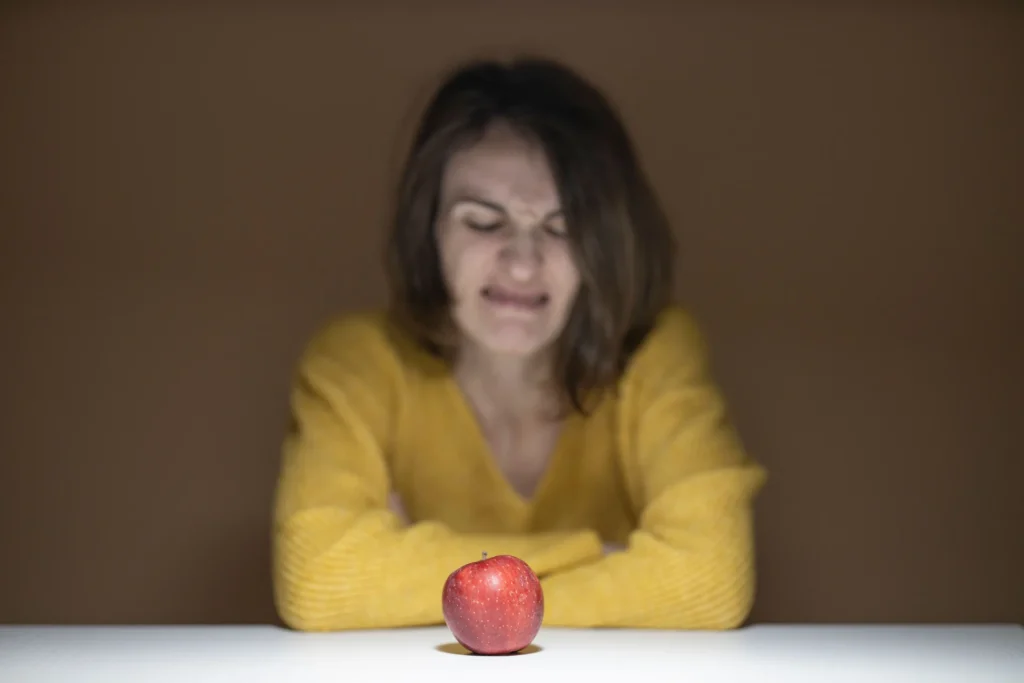 Una donna con un maglione giallo guarda con disgusto una mela rossa appoggiata sul tavolo