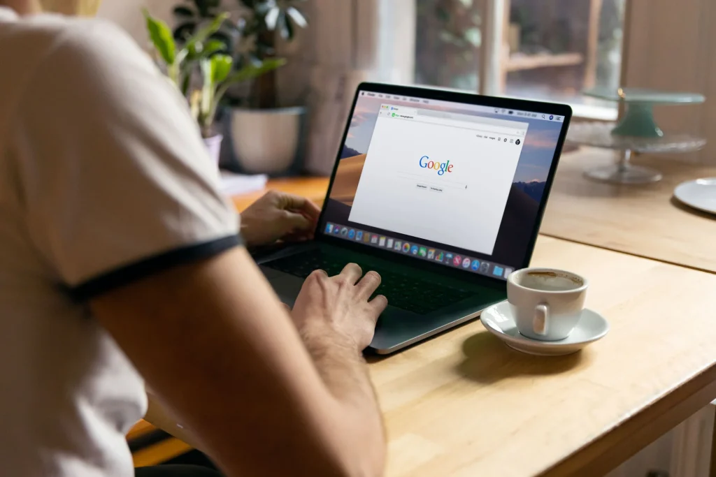 Una persona di spalle digita su un portatile con la pagina di ricerca di Google aperta, accanto a una tazza di caffè